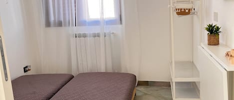 A bedroom with two beds, a window with sheer curtains, a radiator, a white metal shelf with baskets, and a small potted plant.
