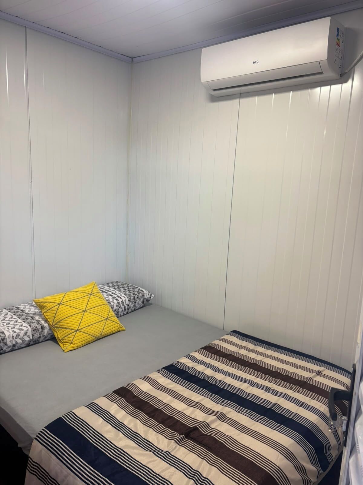A small, single-bed room with a plain white wall, a window, and an air conditioning unit mounted on the wall.