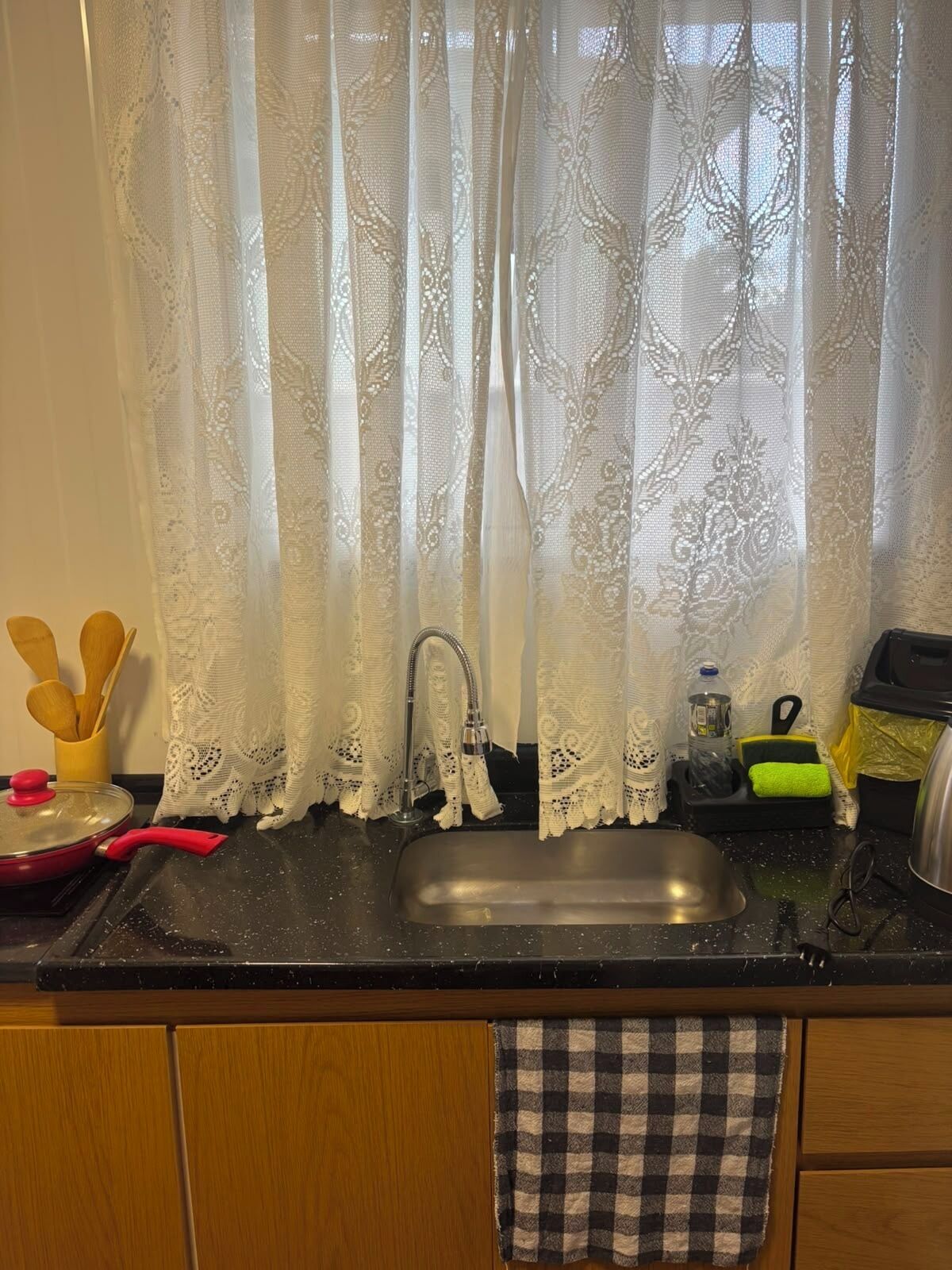 A kitchen with a sink, a faucet, a red pot, and a checkered towel hanging on the cabinet door.