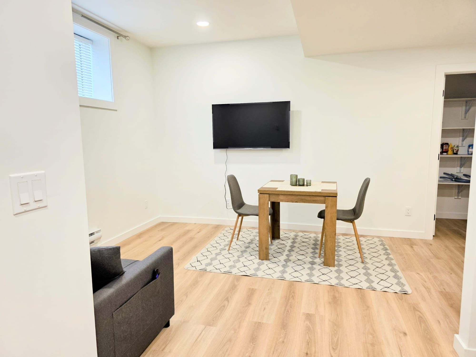 Una sala de estar moderna con una mesa de comedor de madera, dos sillas, un televisor de pantalla plana en la pared y un sofá gris.
