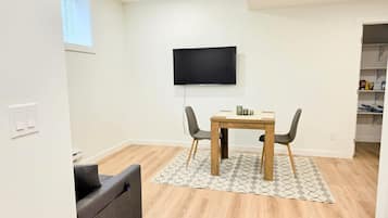 Una sala de estar moderna con una mesa de comedor de madera, dos sillas, un televisor de pantalla plana en la pared y un sofá gris.