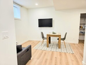 Una sala de estar moderna con una mesa de comedor de madera, dos sillas, un televisor de pantalla plana en la pared y un sofá gris.