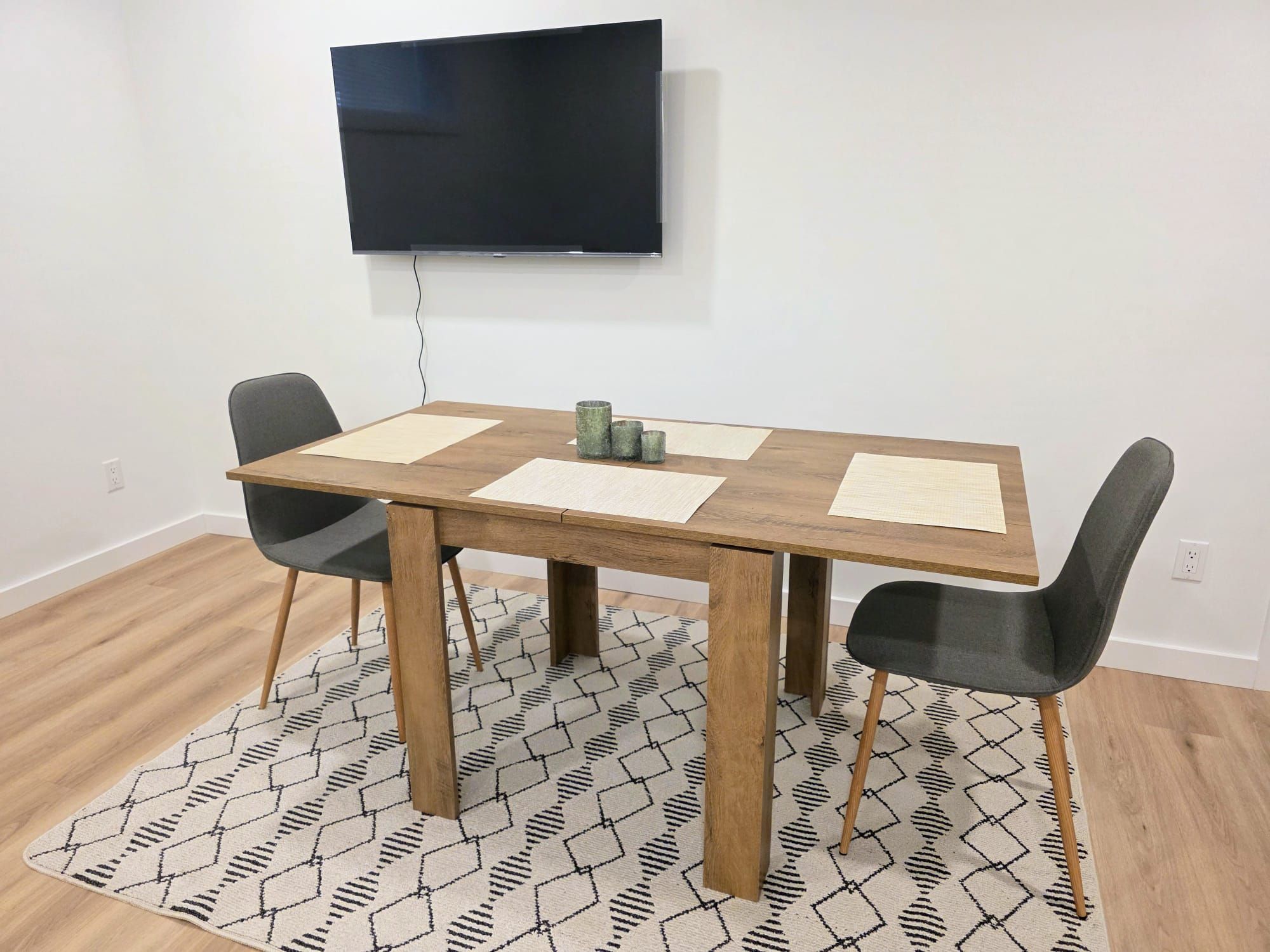 A dining table with four chairs, a television mounted on the wall, and a patterned rug on the floor.