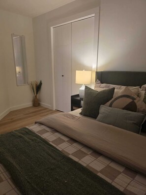 A bedroom with a bed, a nightstand, a lamp, a mirror, and a door. - Snuggle Inn: Cozy 1BR Suite in Rosewood | Full Kitchen & Free Parking (Saskatoon)