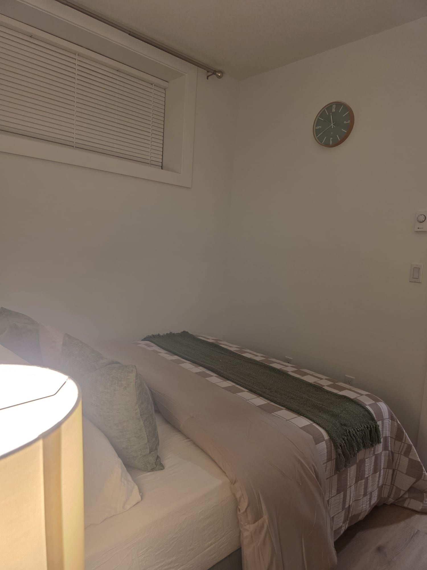 Een kleine eenpersoonskamer met raam, wandklok en bedlampje.