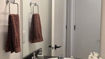 Ein Badezimmer mit einem weißen Waschbecken, einem Spiegel, einem Handtuchhalter mit zwei Handtüchern, einer Dove Seife, einer Tube Crest Zahnpasta und einer Topfpflanze.