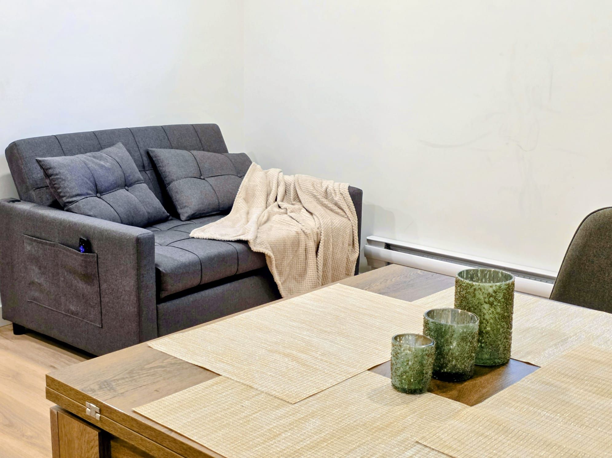 Een moderne woonkamer met een grijze bank, een beige plaid en een houten eettafel met glazen servies.