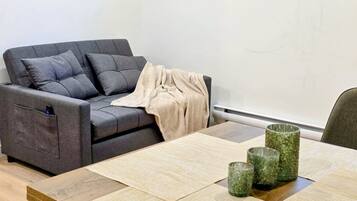 Een moderne woonkamer met een grijze bank, een beige plaid en een houten eettafel met glazen servies.
