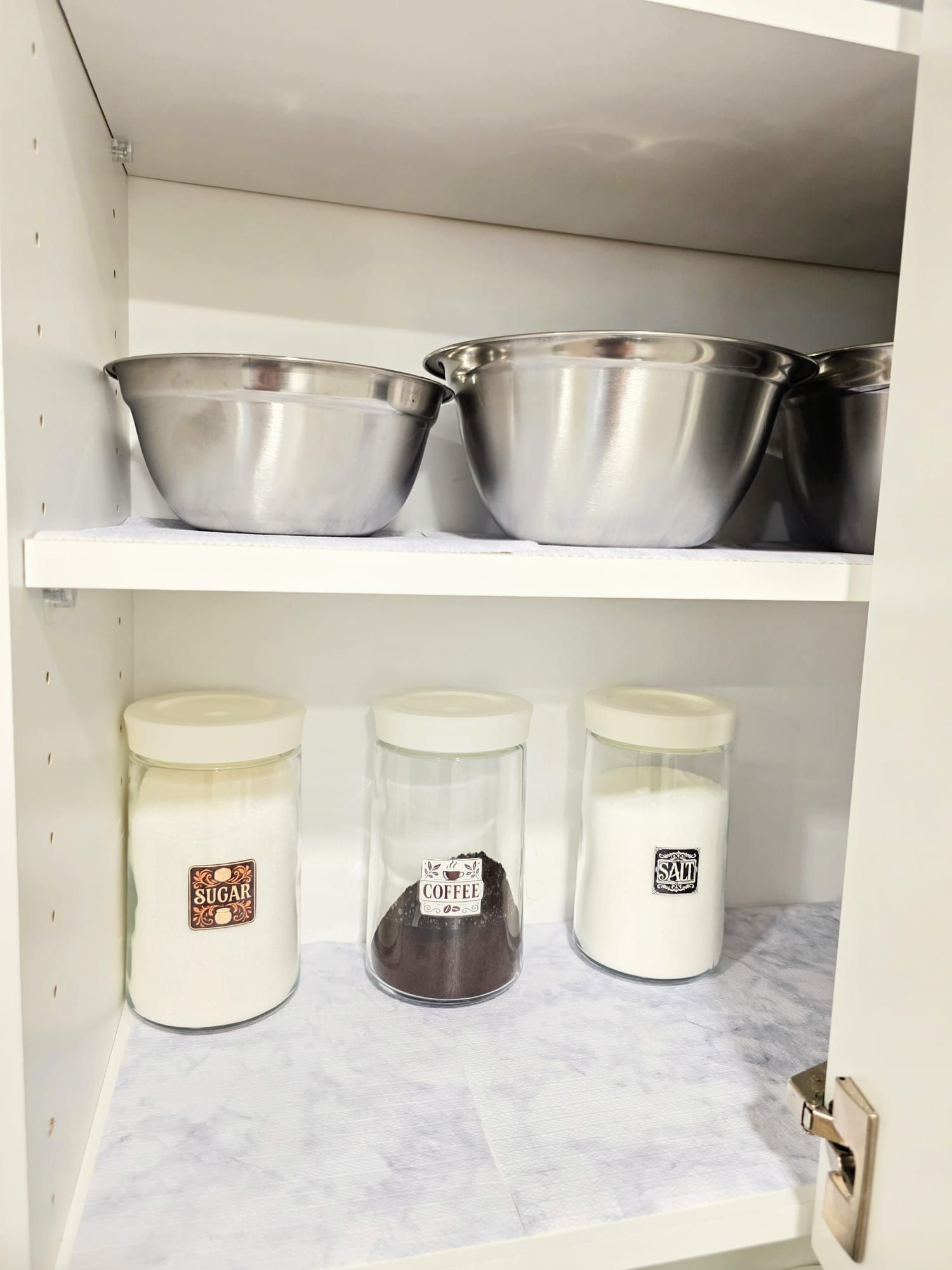A kitchen shelf with three metal bowls and three glass jars with labels.