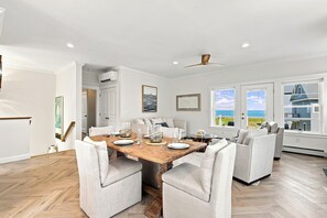 Een eetruimte met een houten tafel, witte fauteuils en uitzicht op de zee door grote ramen.