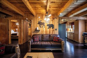 Ein gemütliches Blockhaus-Interieur mit Holzbalkendecke, einer Ledersofa mit gemusterten Kissen, einem Deckenventilator und einer Küchenzeile mit Holzausbau.