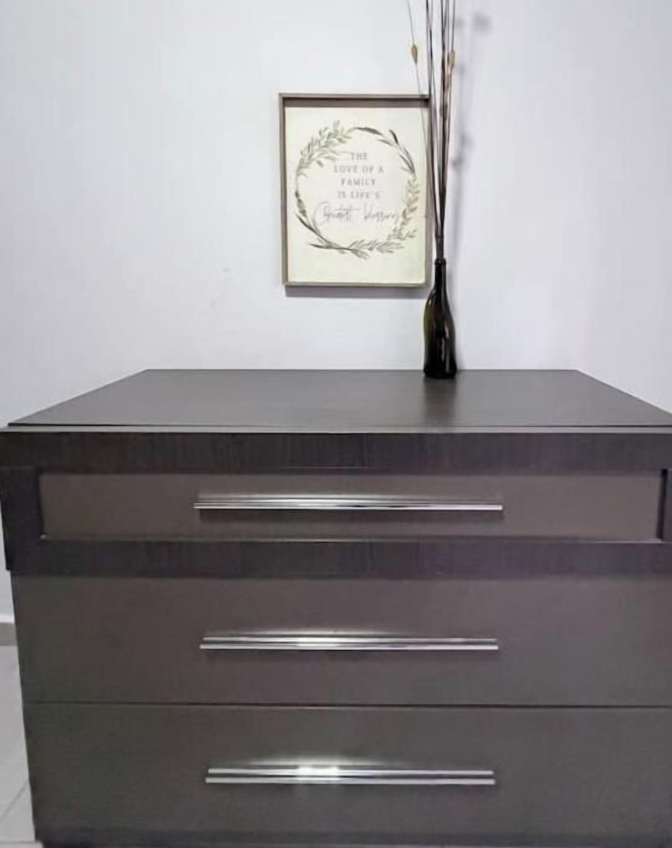 A dark-colored dresser with three drawers and a framed quote on the wall above it.