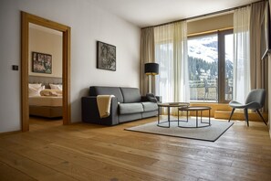 Un salon moderne avec un canapé, une table basse et un fauteuil. On aperçoit un lit avec une tête de lit à travers une porte ouverte.