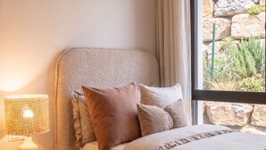 Een slaapkamer met een bed, kussens, een nachtkastje, een lamp en een raam met uitzicht op rotsen en begroeiing.