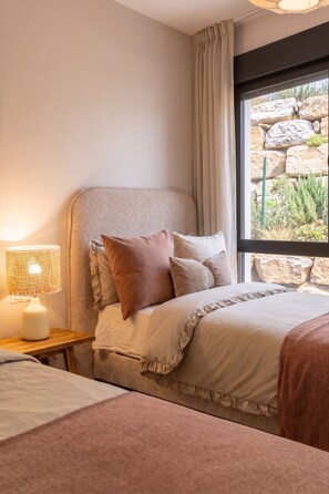 Ein Schlafzimmer mit Bett, Kissen, einem Nachttisch, einer Lampe und einem Fenster mit Blick auf Felsen und Grün.