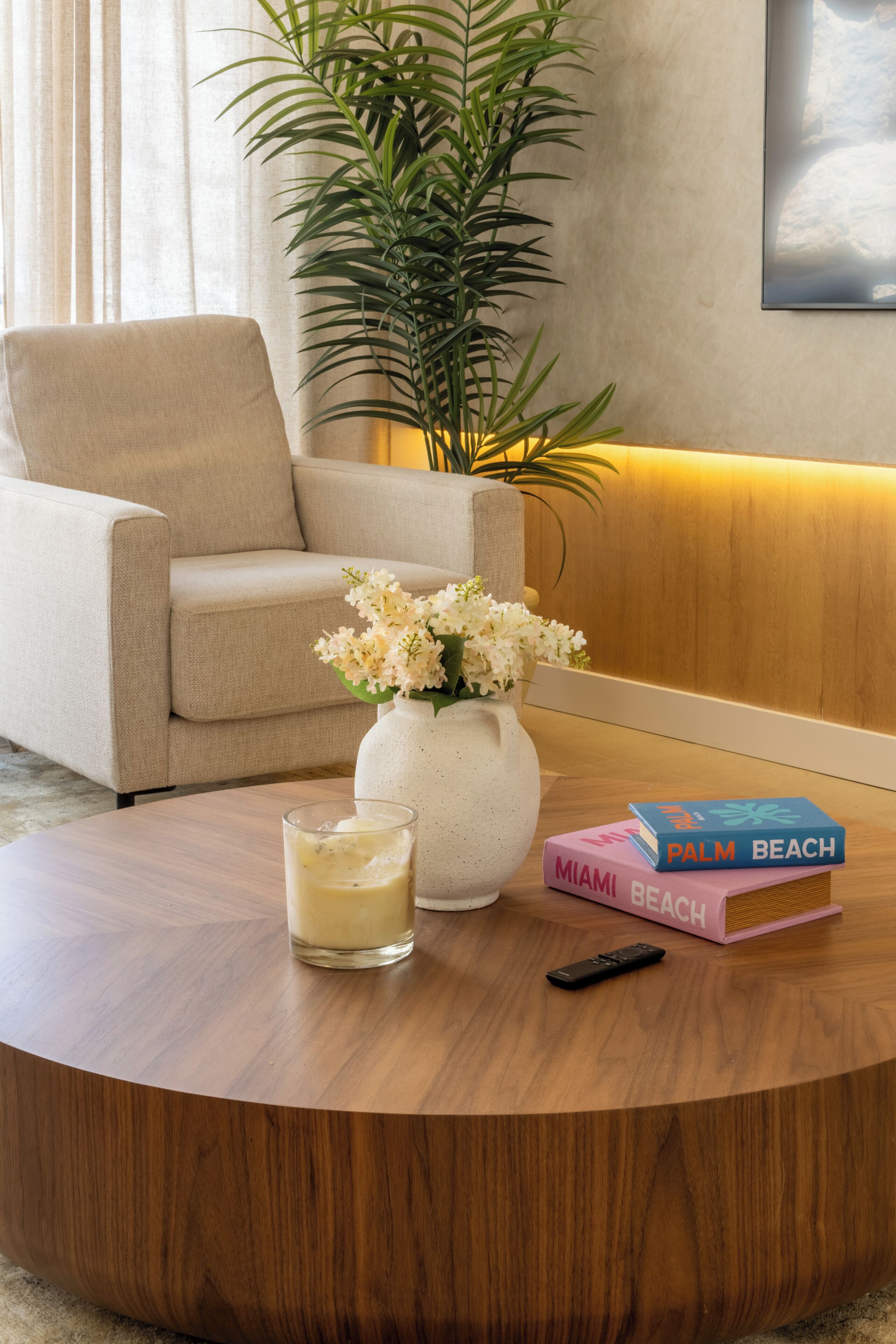 Une table basse ronde en bois, sur laquelle se trouvent un verre de lait, un vase blanc rempli de fleurs et un livre intitulé « MIAMI BEACH ».