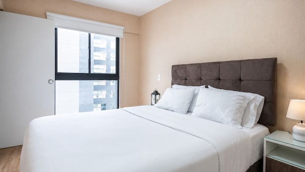 Ein Hotelzimmer mit einem Bett, einem Nachttisch, einer Lampe und Blick auf die Stadt durch ein Fenster.