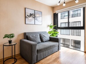 En moderne stue med en grå sofa, et rundt bord med en plante og et stort vindue med persienner.