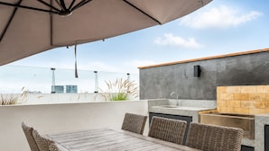 Eine Außenpatio mit Holztisch und Korbstühlen, eine integrierte Küche mit Spüle und Arbeitsplatte, und Blick auf die Skyline der Stadt.