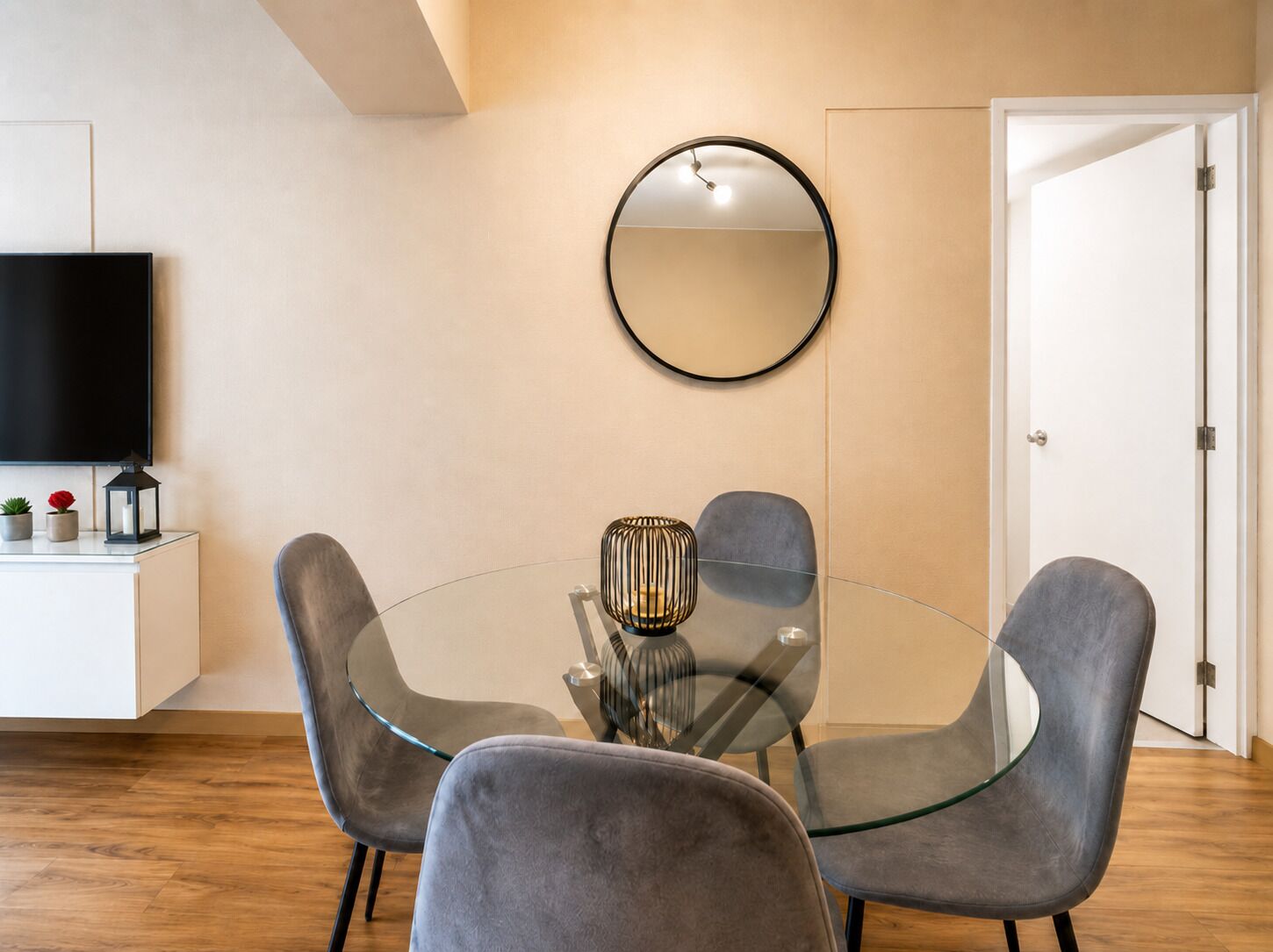 A modern dining area with a glass table surrounded by grey chairs, a round mirror on the wall, and a flat-screen TV on a white cabinet.