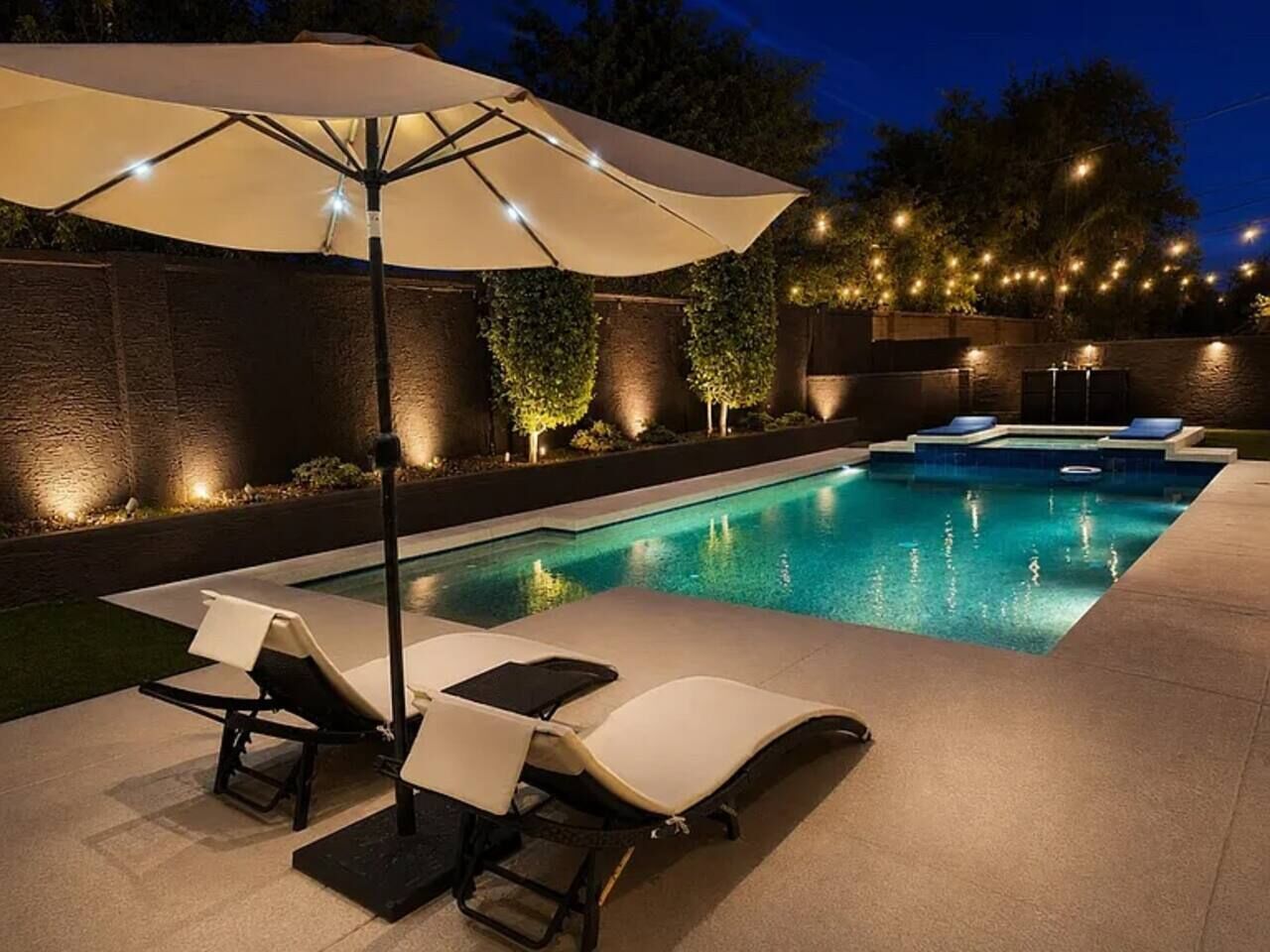 A swimming pool with lounge chairs and an umbrella, surrounded by a brick wall and illuminated by string lights.