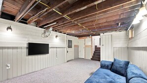 An attic room with a television mounted on the wall, a blue sofa, and a staircase leading to another level. - The Copper & Pine Cottage (Anaconda)