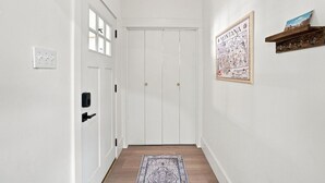A hallway with a white door, a wall-mounted light switch, a framed map, and a wooden shelf with hooks. - The Copper & Pine Cottage (Anaconda)