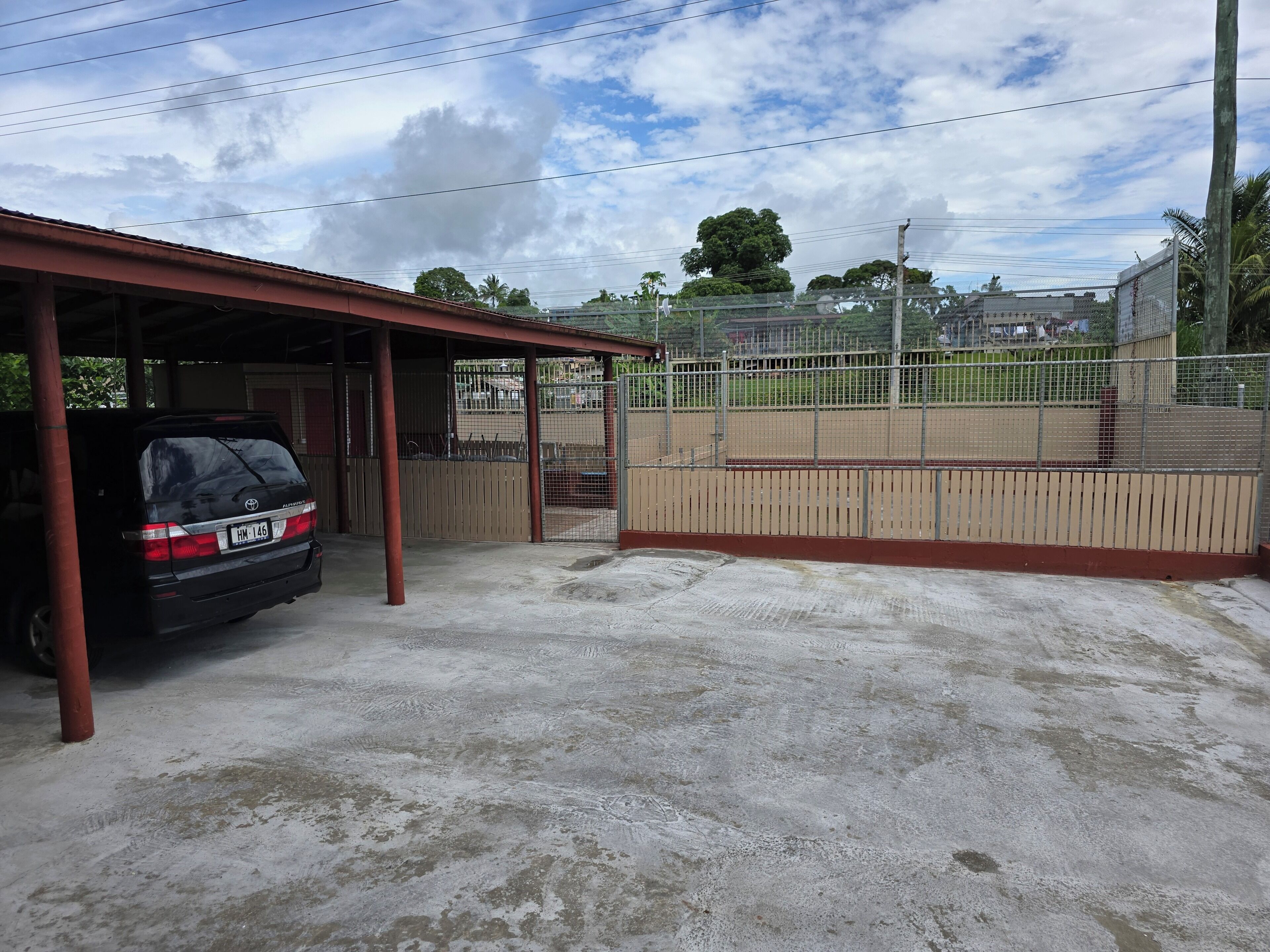 Un área de estacionamiento techada con un coche aparcado en su interior, un campo deportivo cercado y un edificio con techo rojo al fondo.