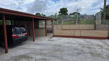Un área de estacionamiento techada con un coche aparcado en su interior, un campo deportivo cercado y un edificio con techo rojo al fondo.