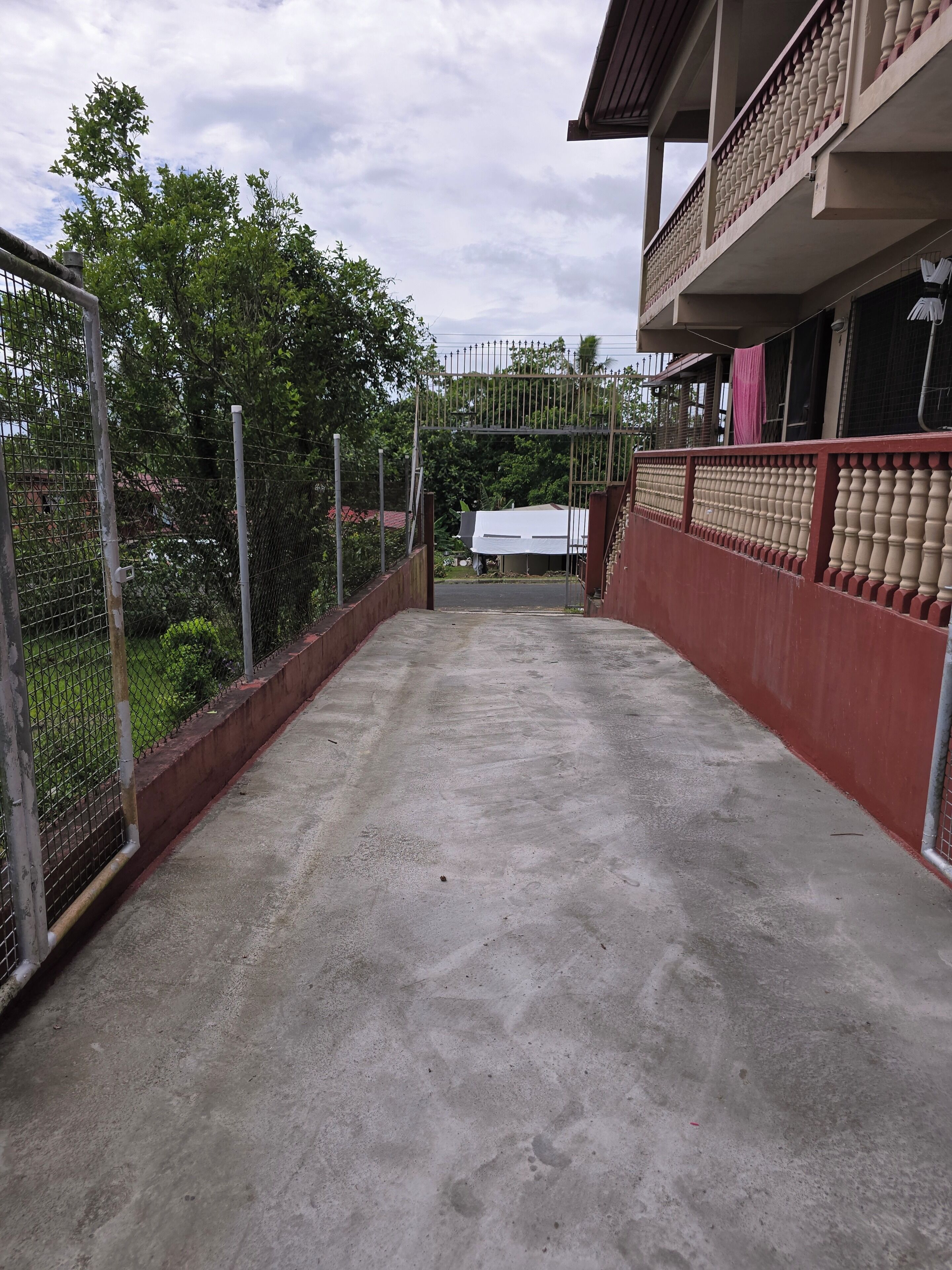 Un camino de concreto lleva a una entrada con portón, baranda roja y un edificio blanco al fondo.