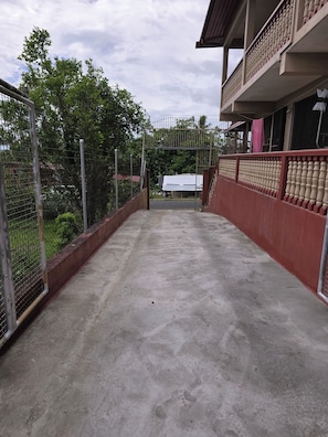 Un camino de concreto lleva a una entrada con portón, baranda roja y un edificio blanco al fondo.