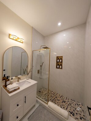 Un baño moderno con ducha de vidrio, un espejo grande y un tocador con lavamanos y artículos de aseo.