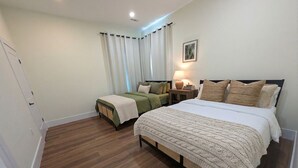 Ein Schlafzimmer mit einem großen Bett, einem kleineren Bett, einem Nachttisch mit Lampe und einem Fenster mit Vorhängen.