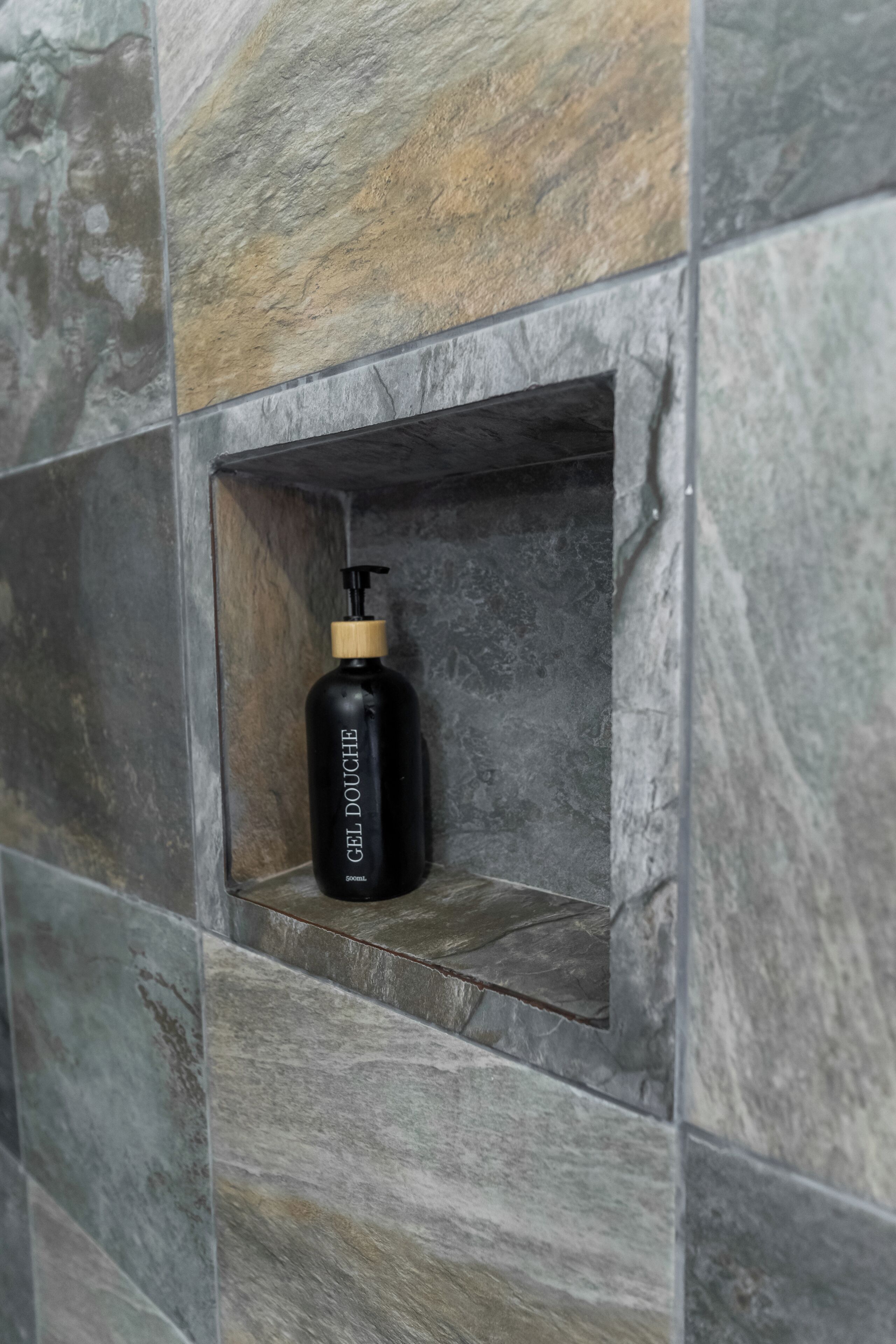 A black bottle with a pump dispenser labeled 'GEL DOUCHE' in a recessed stone wall alcove.