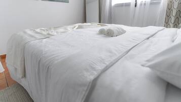 A neatly made bed with white linens and two rolled towels, a mirror on the wall, and a window with blinds.