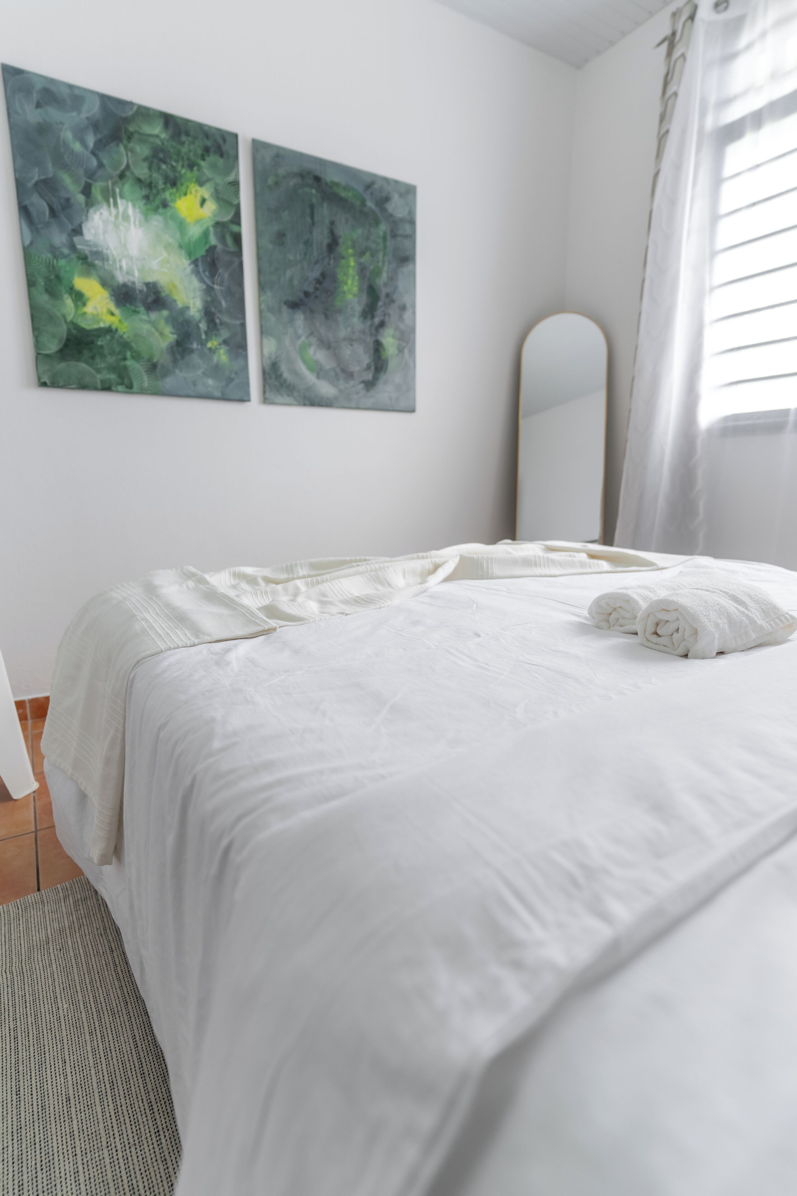 A neatly made bed with white linens and two rolled towels, a large mirror, and two abstract paintings on the wall.