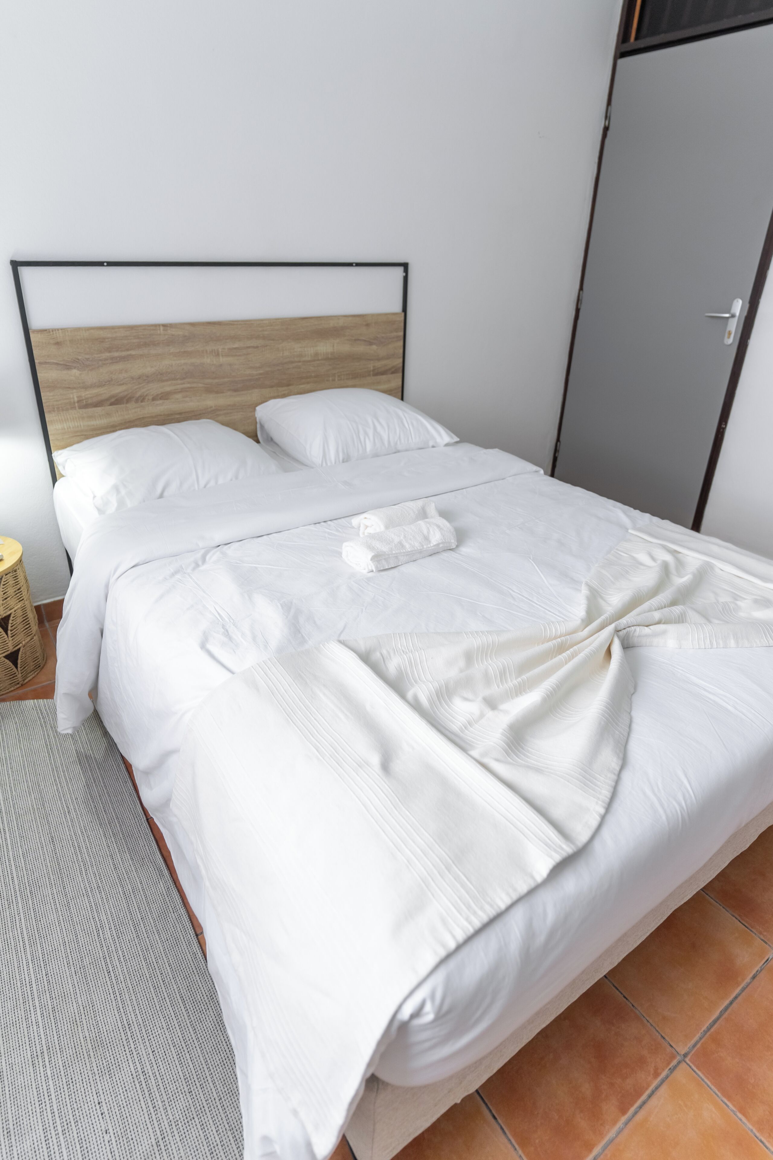 Un lit bien fait, avec du linge de lit blanc et une tête de lit en bois, une armoire grise et une petite table d’appoint ronde en osier.