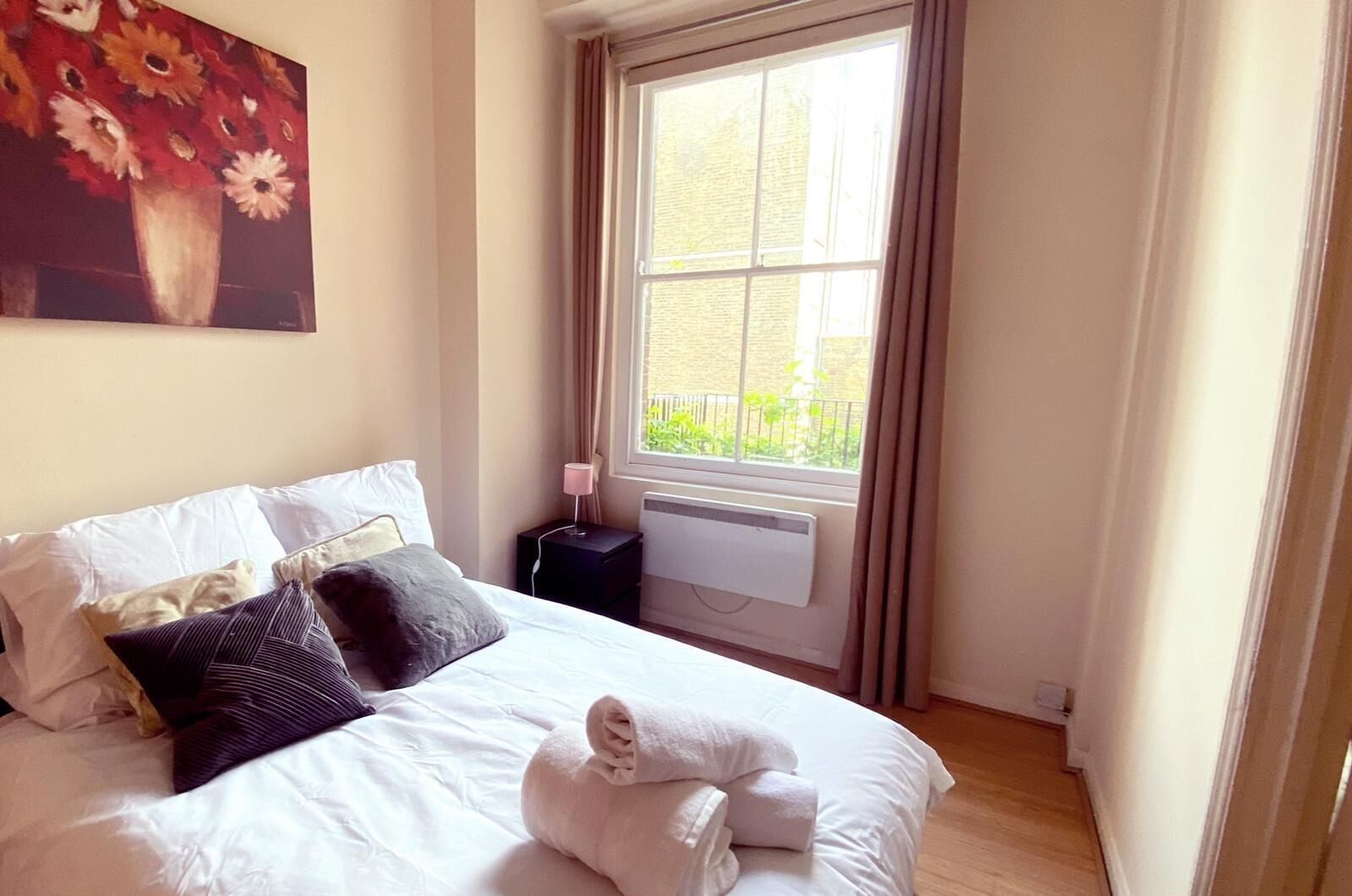 A neatly made bed with white linens and a rolled towel, a bedside lamp, and a window with curtains.