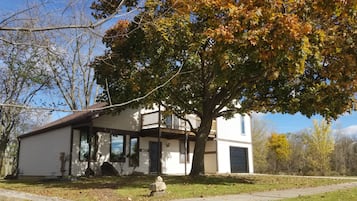 Una casa de dos pisos con un porche, un árbol grande al frente y un patio cubierto de césped.