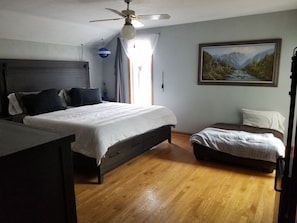 Una camera da letto con un letto, una scrivania, una sedia, una ventola a soffitto, una finestra con tende e un quadro appeso al muro.