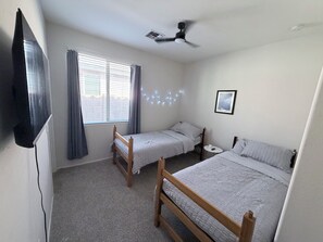 Un dormitorio con dos camas individuales, un ventilador de techo, un ventanal con cortinas y una mesita de noche pequeña.