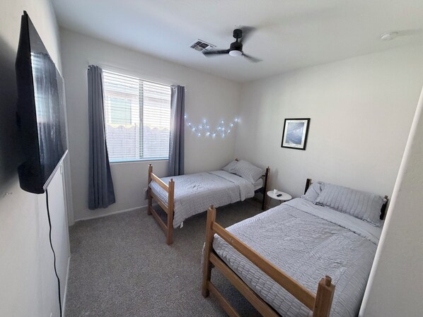 A bedroom with two single beds, a ceiling fan, a window with curtains, and a small bedside table.