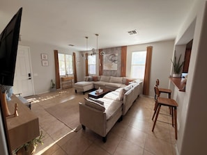 Una sala moderna con un sofá modular, una mesa de centro y un televisor. Hay una mesita auxiliar de madera con una planta, y una ventana con cortinas.