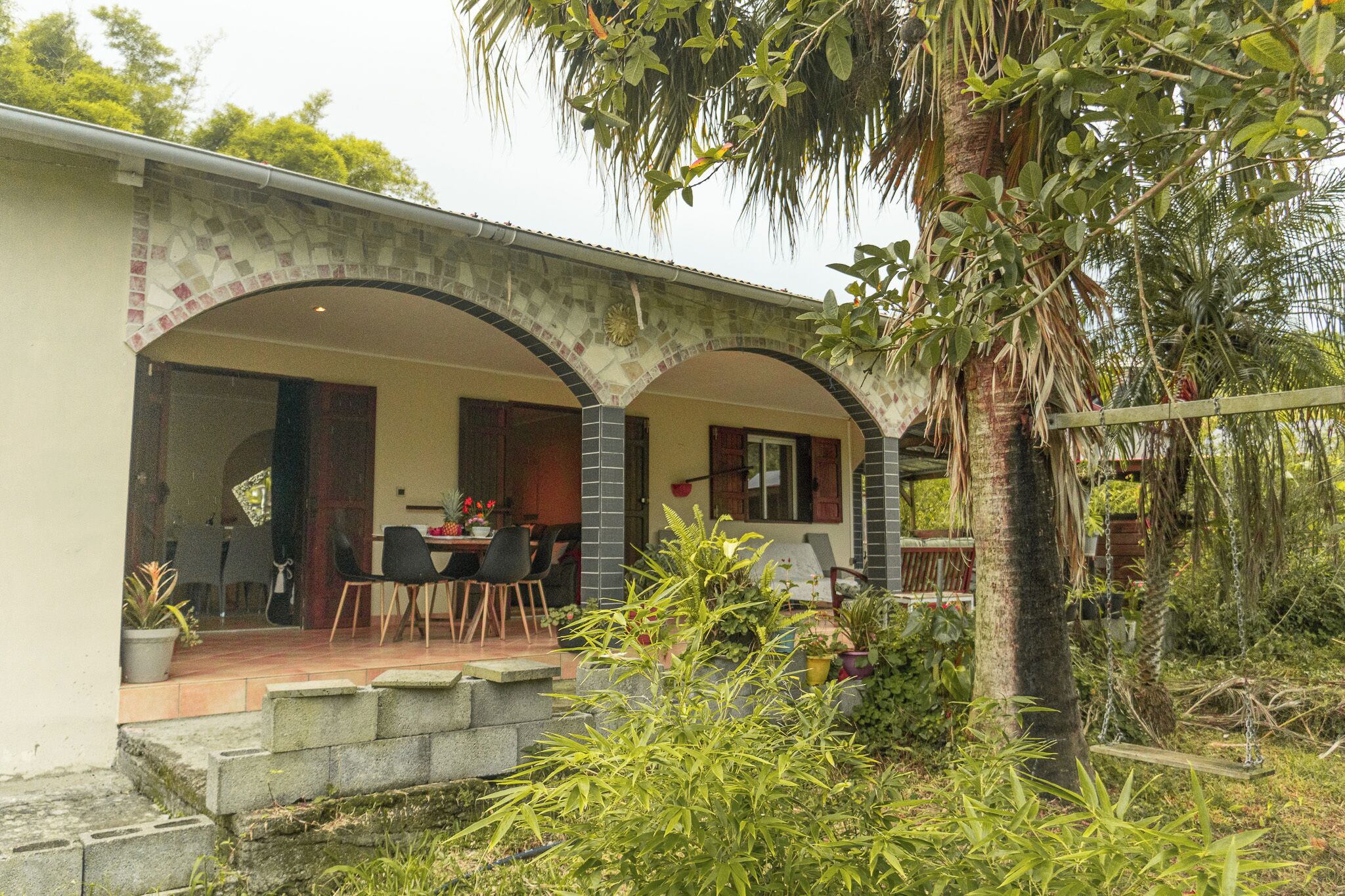 A covered outdoor dining area with a table and chairs, surrounded by greenery and a palm tree.