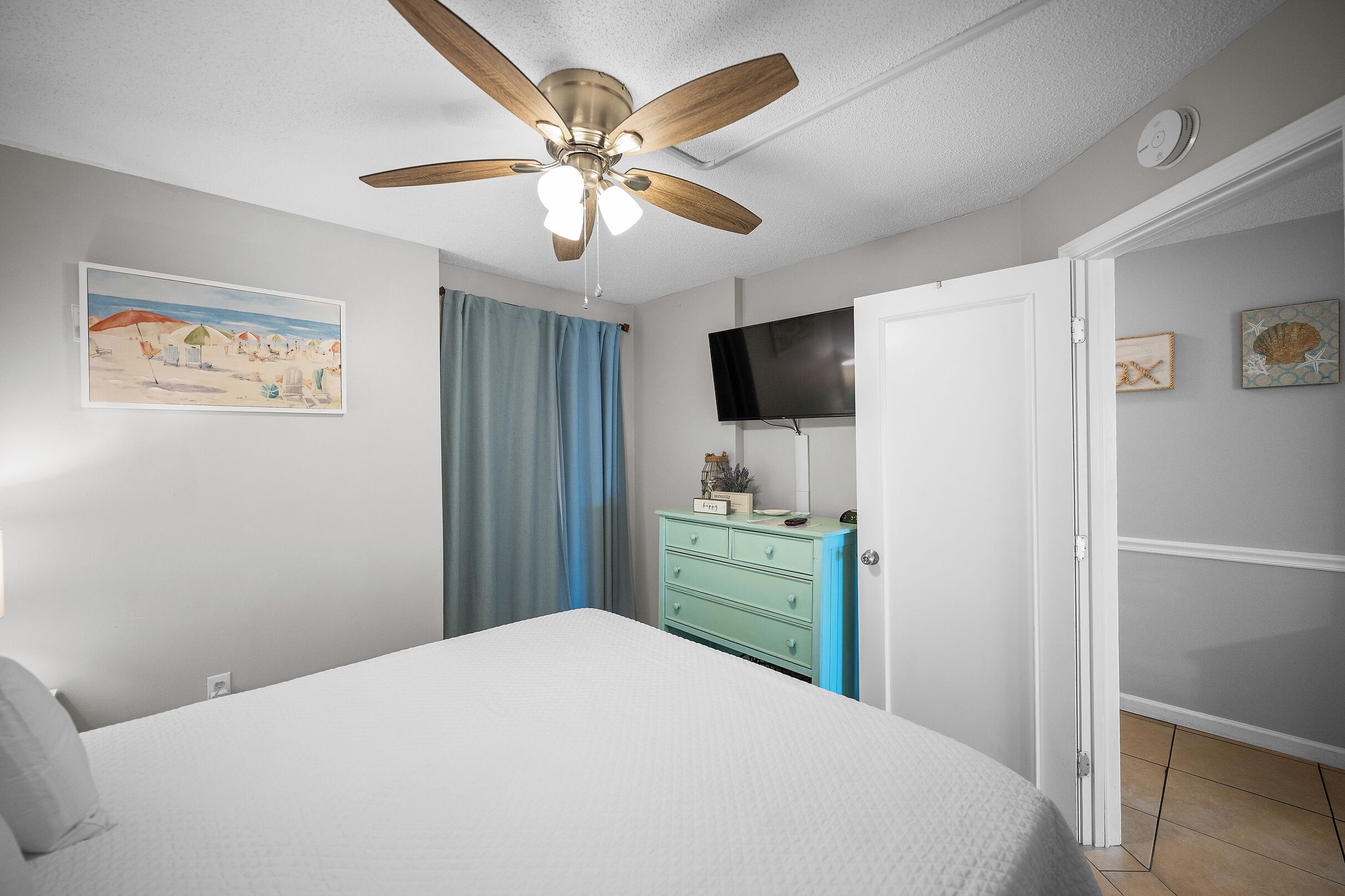 A bedroom with a bed, a ceiling fan, a television, a dresser, and a door.