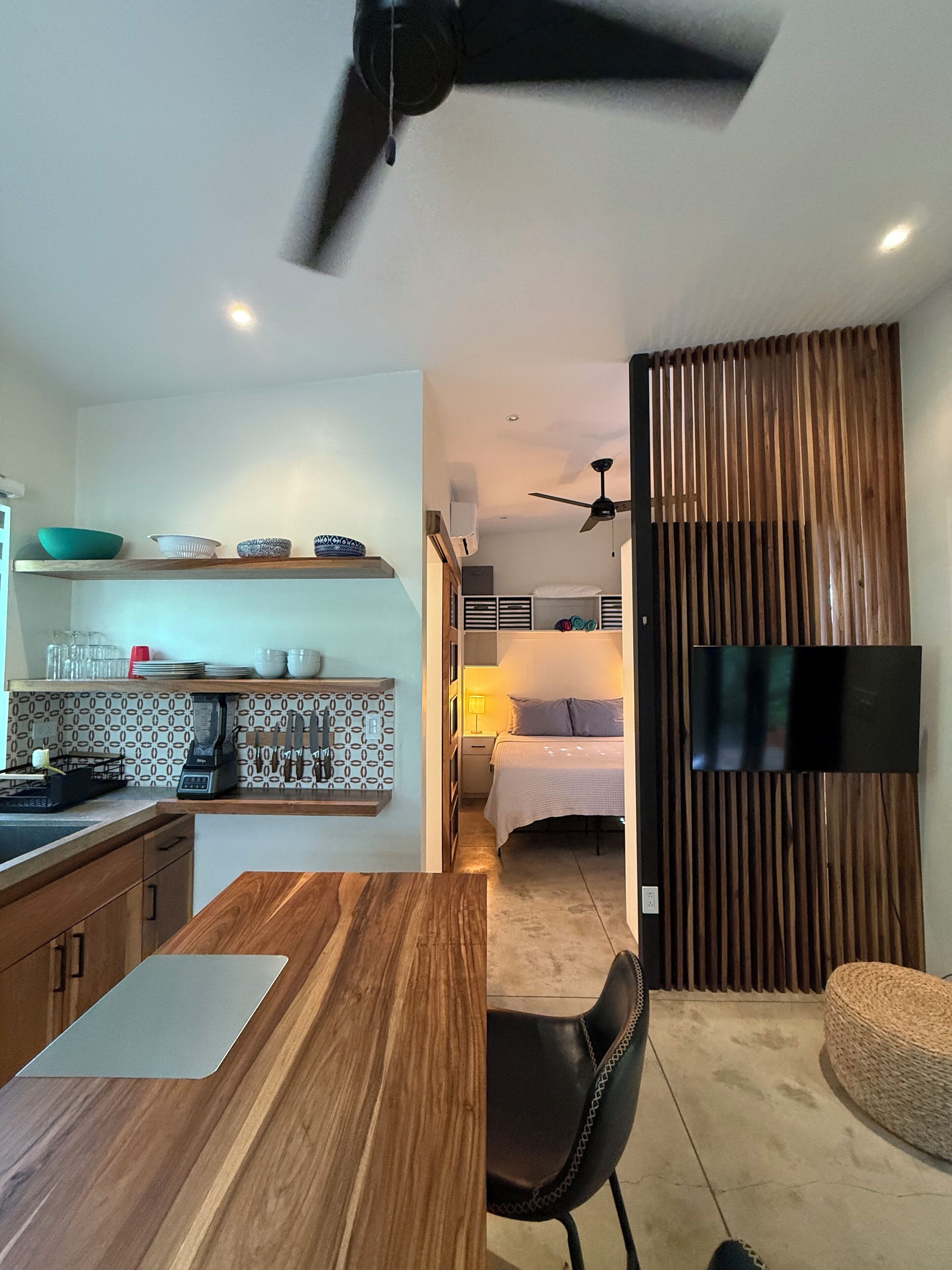 Une cuisine moderne avec un plan de travail en bois, un coin repas avec une table et des chaises, une chambre avec un lit et une télévision, et un salon avec un canapé et un pouf en osier.
