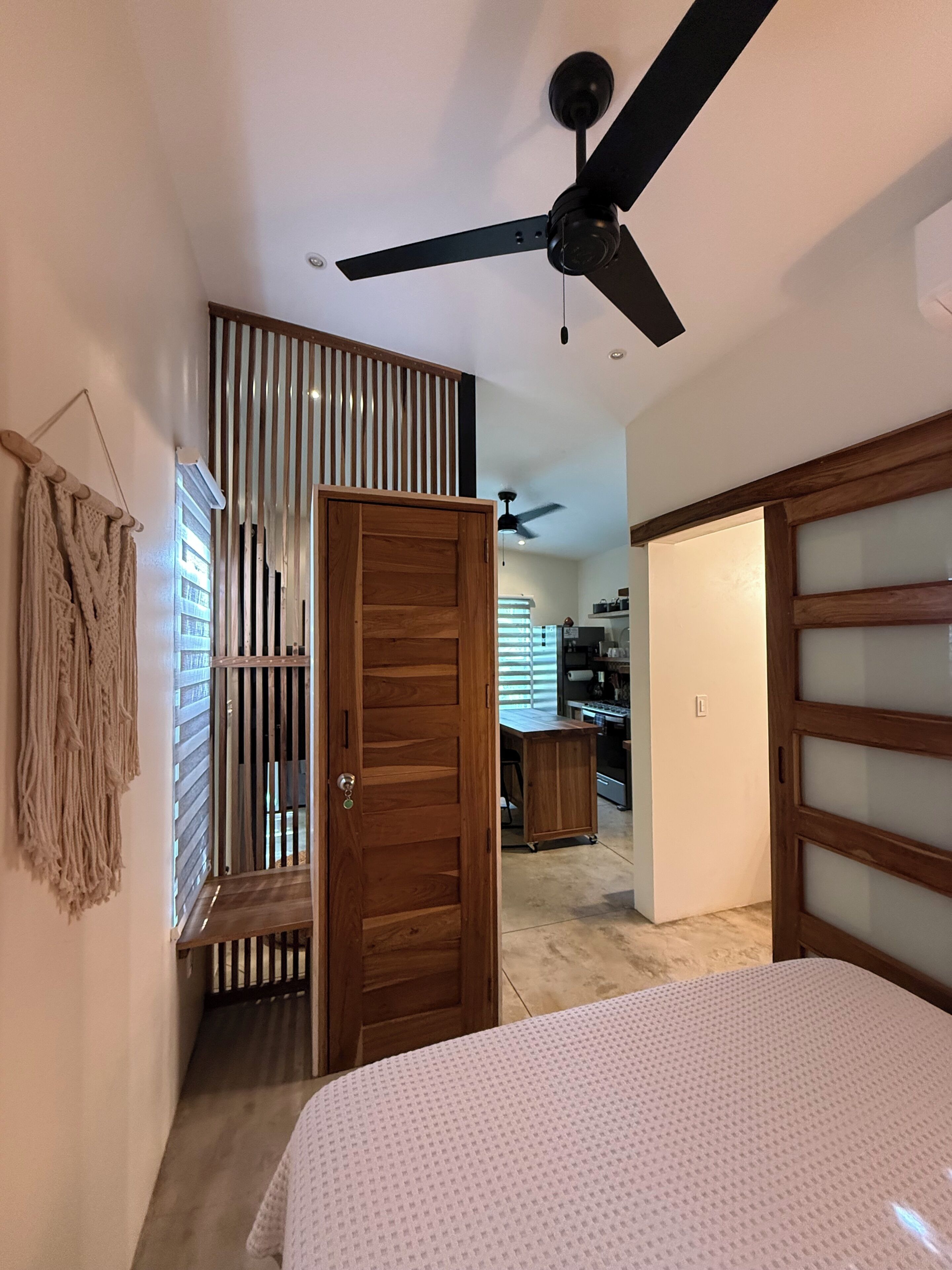 A bedroom with a bed, a ceiling fan, a wardrobe, and a kitchen area in the background.