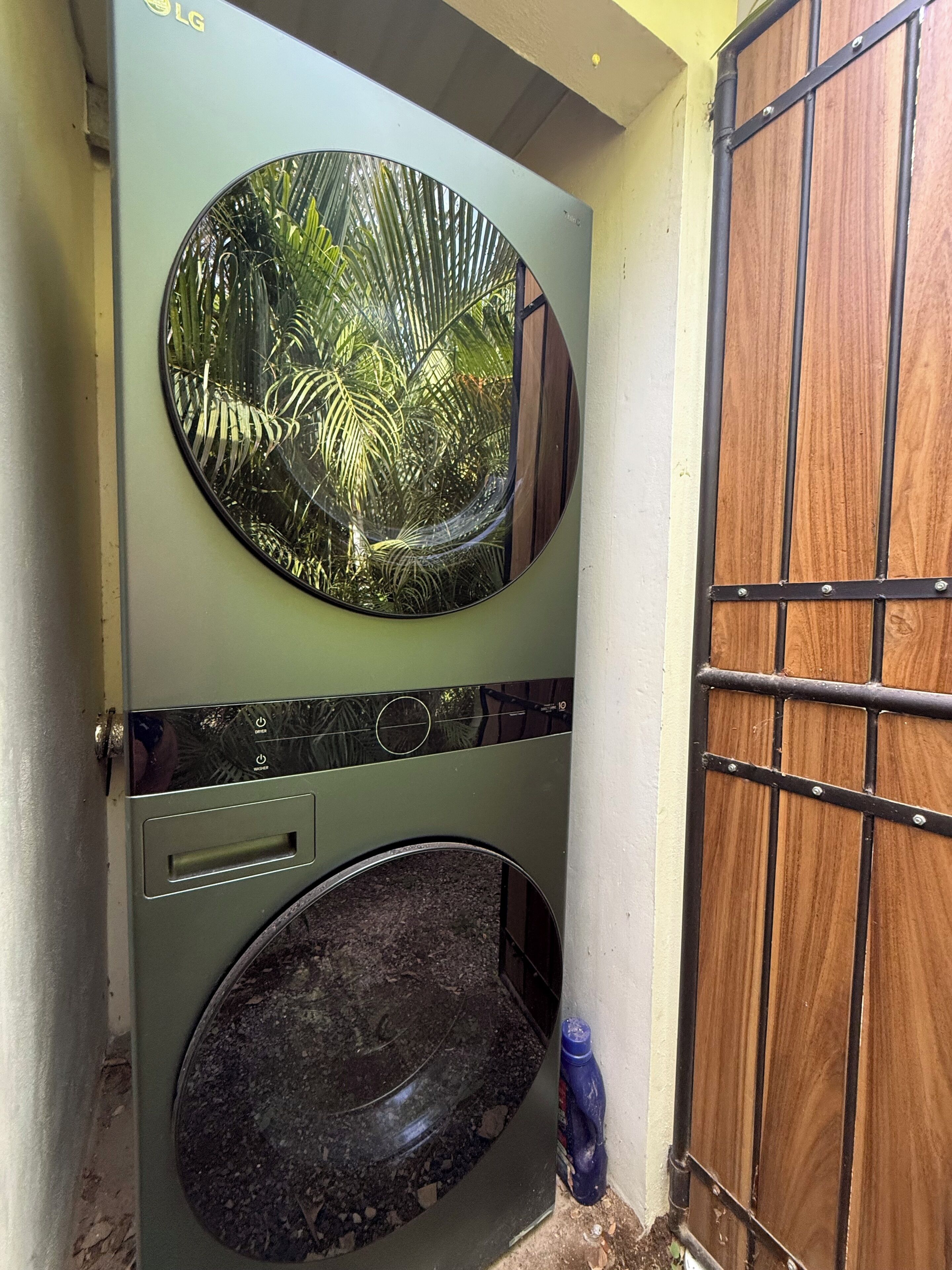 A wall-mounted washing machine with a circular window, a metal door with a grid pattern, and a small blue bottle on the floor.