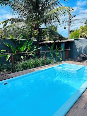 A swimming pool with a wooden deck, surrounded by lush greenery and a small house with a tiled roof. - Pousada Refugá (Porto Seguro)