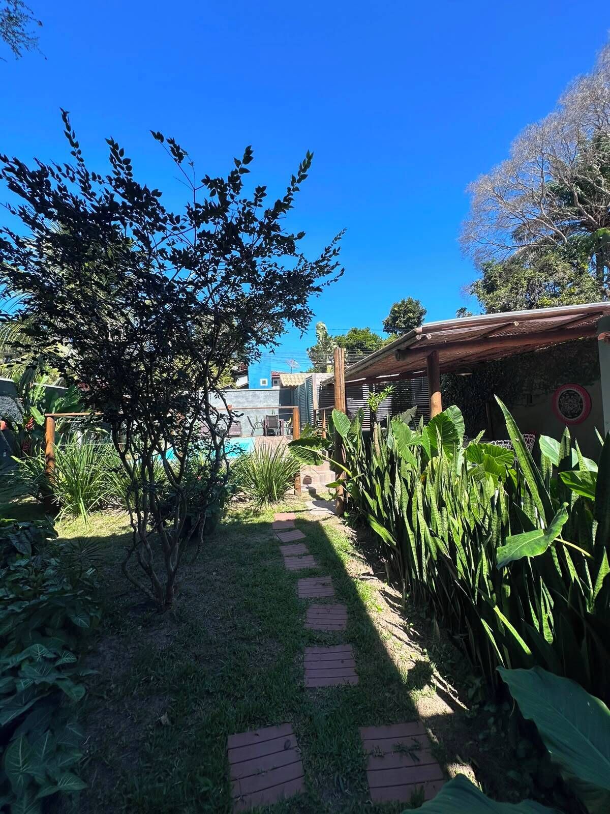 A garden pathway leading to a building with a pool, surrounded by lush greenery and a clear blue sky.